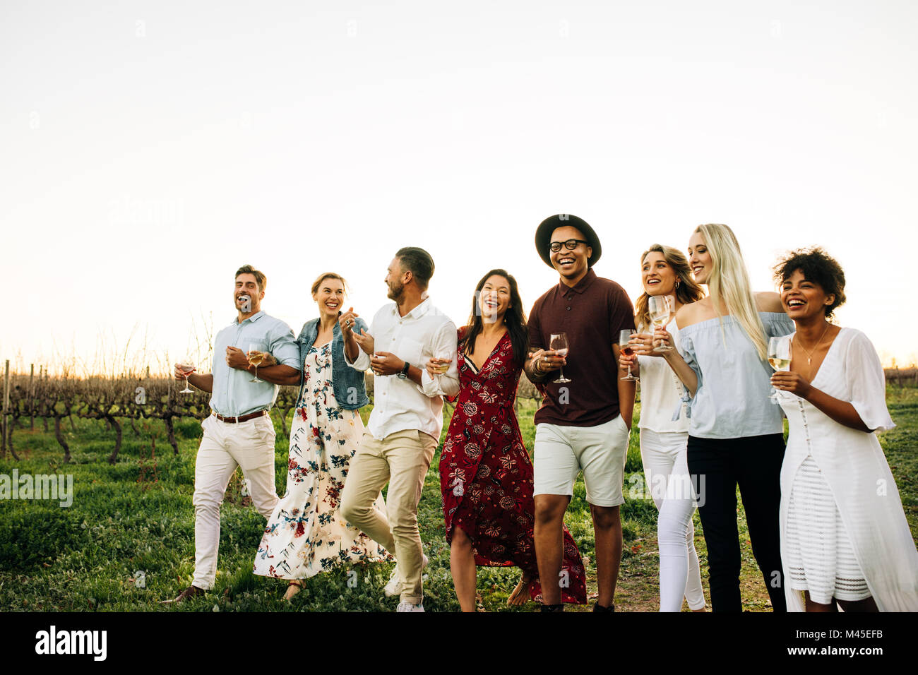 Group of friends enjoying a walk outside in countryside. Multi-ethnic millennials with drink partying outdoors in vineyard. Stock Photo