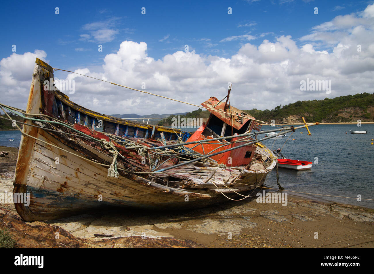 Old fishing boat turned on a side, wrecked and rotting on a rocky shore by the Mira River bank. Bright blue clouded sky. Vila Nova de Milfontes, Portu Stock Photo