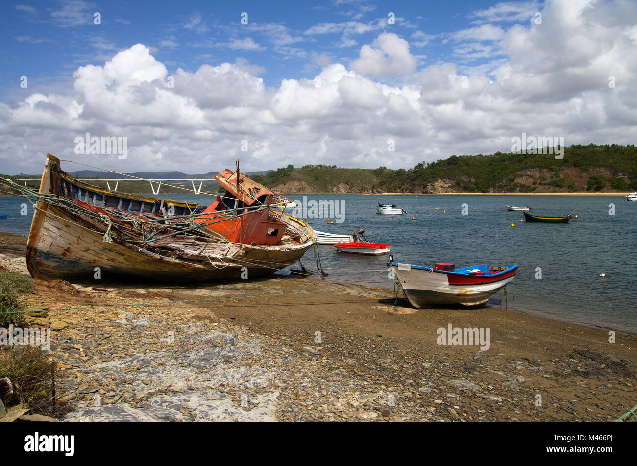 Old trawler fishing boat turned on a side, wrecked and rotting on a rocky shore by the Mira River bank. Smaller boats around. Bright blue clouded sky. Stock Photo