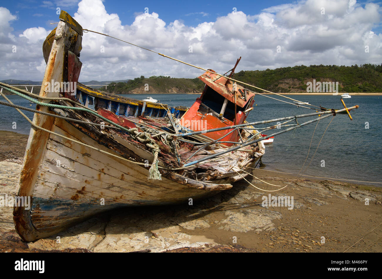 Frontal view of an old fishing boat turned on a side, wrecked and rotting on a rocky bank by the Mira River bank. Bright blue clouded sky. Vila Nova d Stock Photo