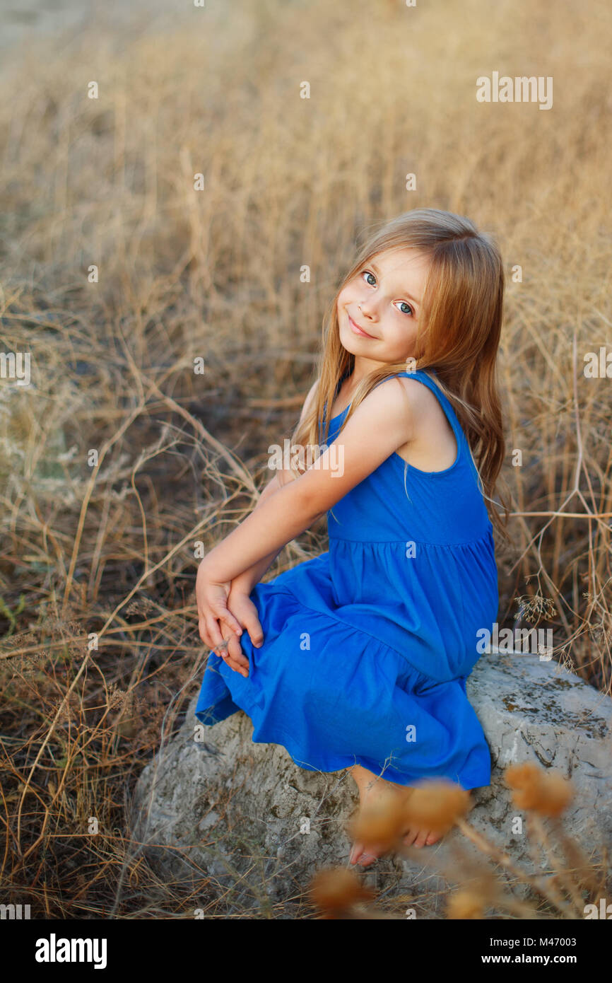 A little girl in a blue dress is sitting on the stones in the middle of ...