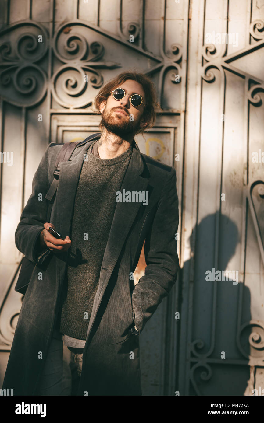Portrait of a confident bearded man in sunglasses and coat holding mobile phone while walking on a city street Stock Photo