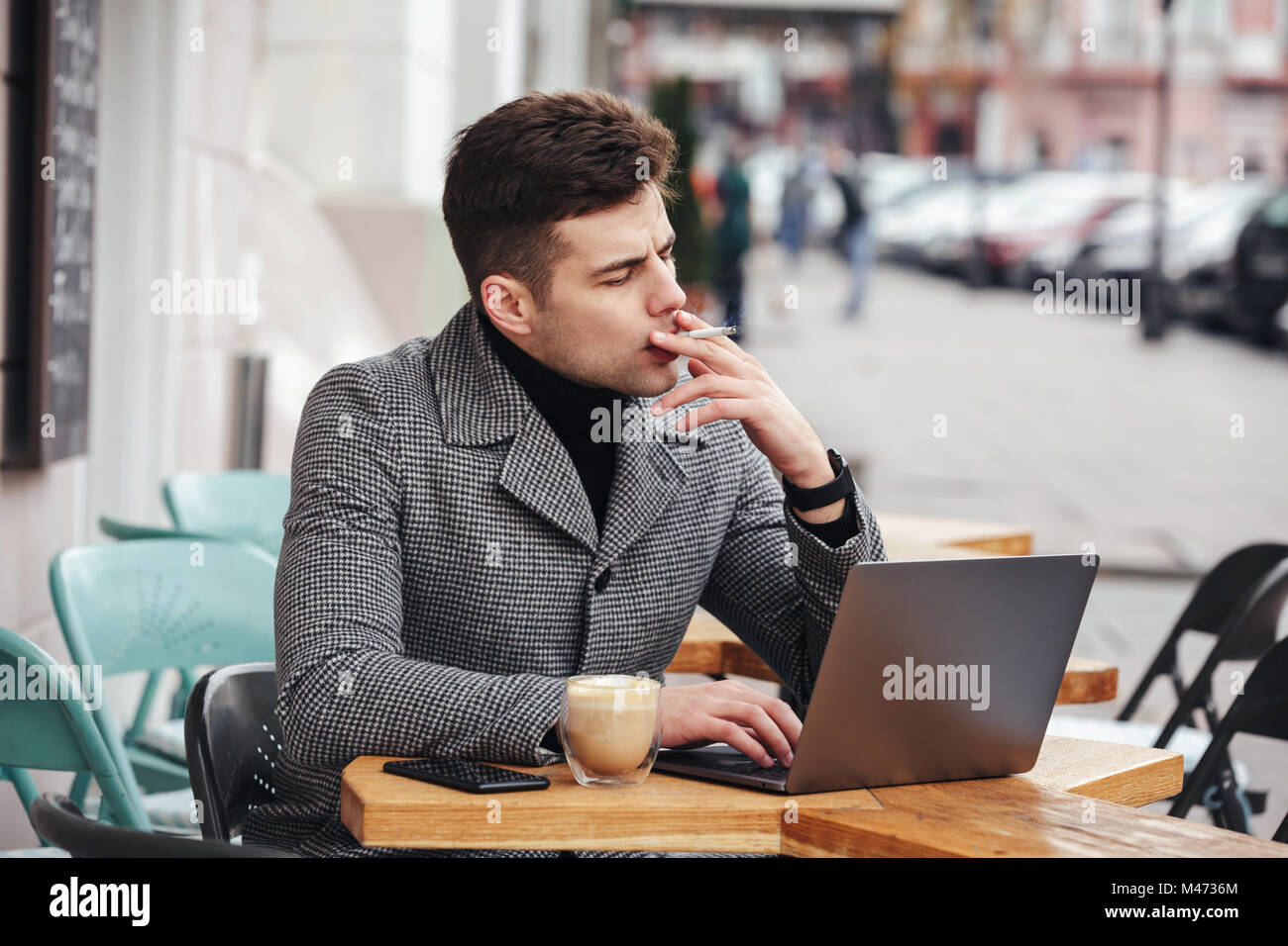 Photo of handsome man in gray coat smoking cigarette and drinking cappuccino while resting in cafe outdoors Stock Photo