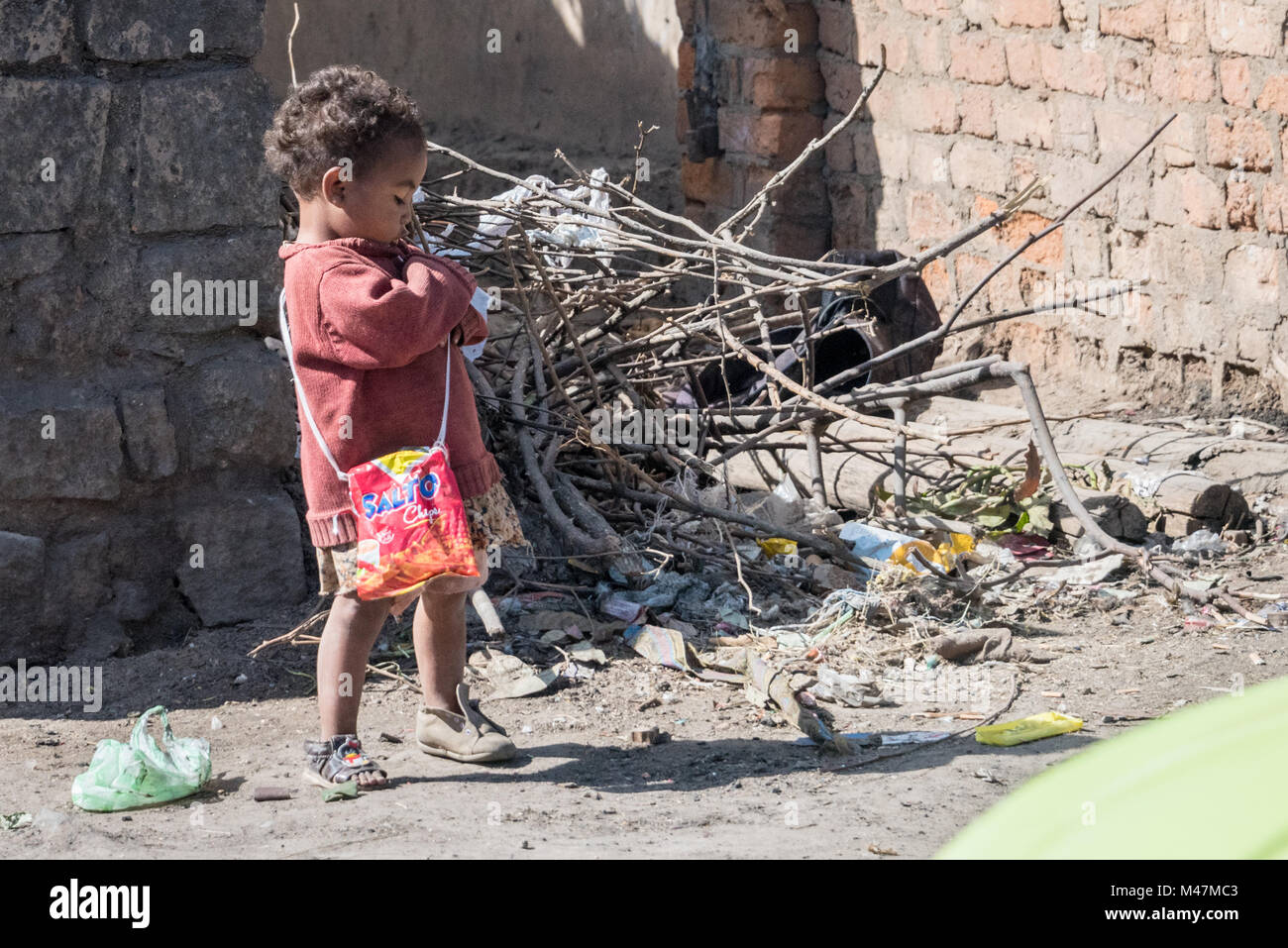 Young Girl, with Odd Shoes, Dirty Coat and Bag in Dust and Rubbish at Roadside, Madagascar Stock Photo