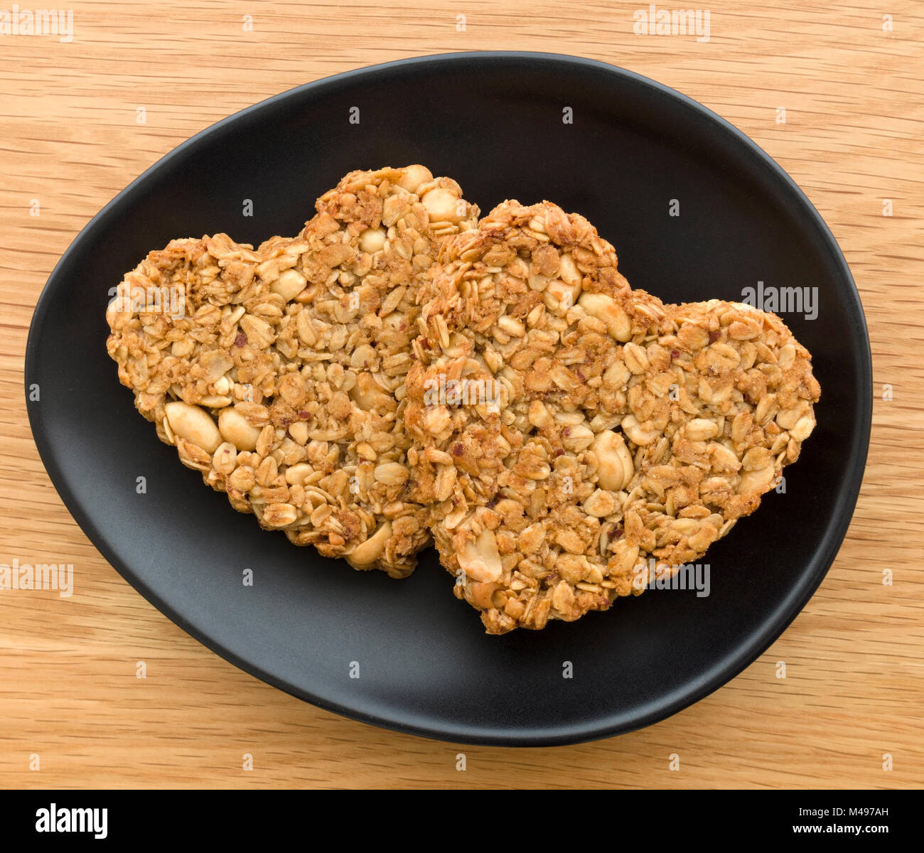 Two home-baked heart shaped peanut and oat flapjack biscuits made for Valentines day on oval black plate on wooden table. Stock Photo