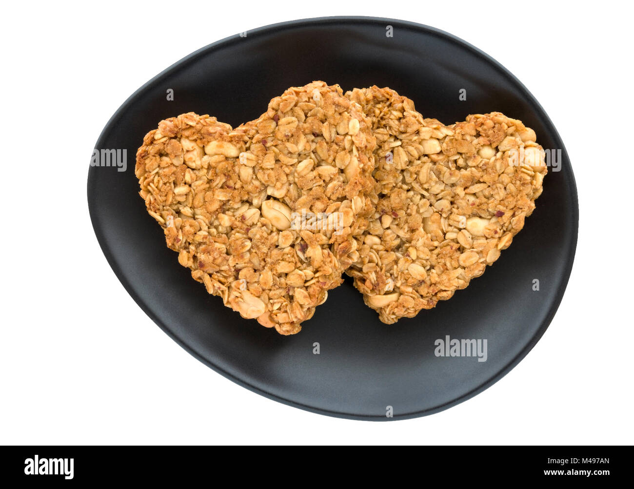 Two home-baked heart shaped peanut and oat flapjack biscuits made for Valentines day on oval black plate and white background. Stock Photo
