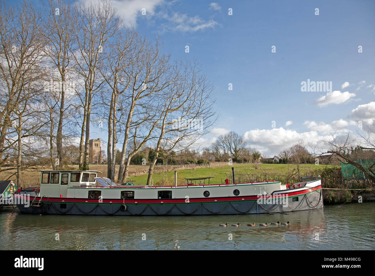 Large barge moored on the Grand Union Canal at Marsworth, Tring, UK., with All Saints Church in the background and mallard ducks in the foreground. Stock Photo
