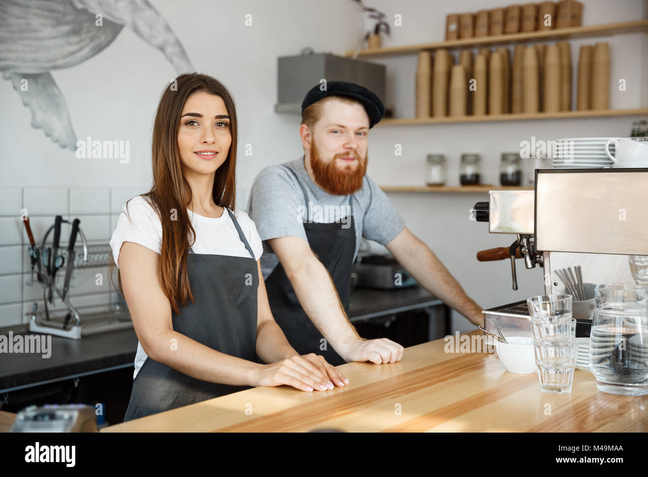 Coffee Business Concept - Positive young bearded man and beautiful attractive lady barista couple in apron looking at camera while standing at bar Counter. Stock Photo