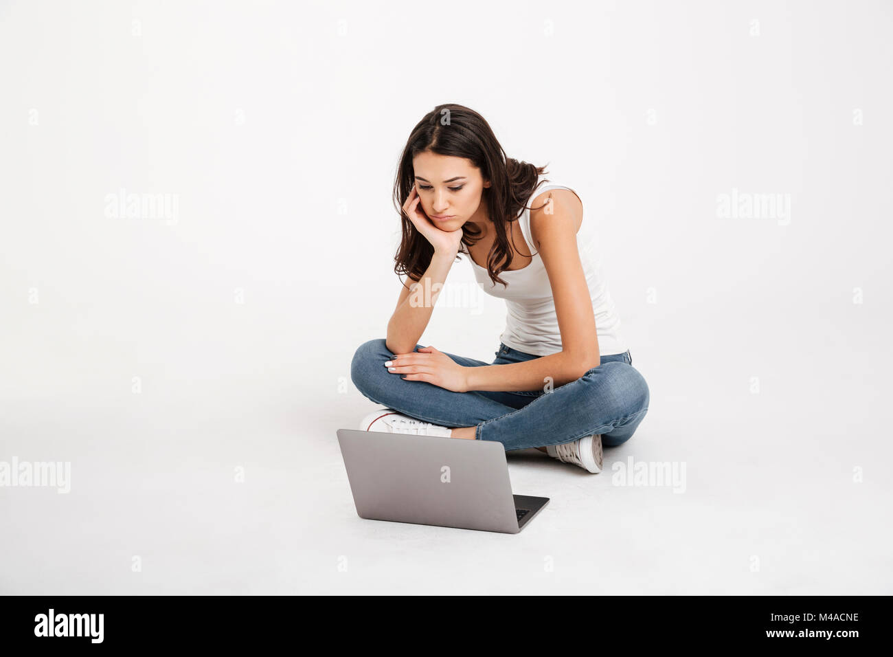 Portrait of a tired girl dressed in tank-top holding laptop while sitting on the floor isolated over white background Stock Photo
