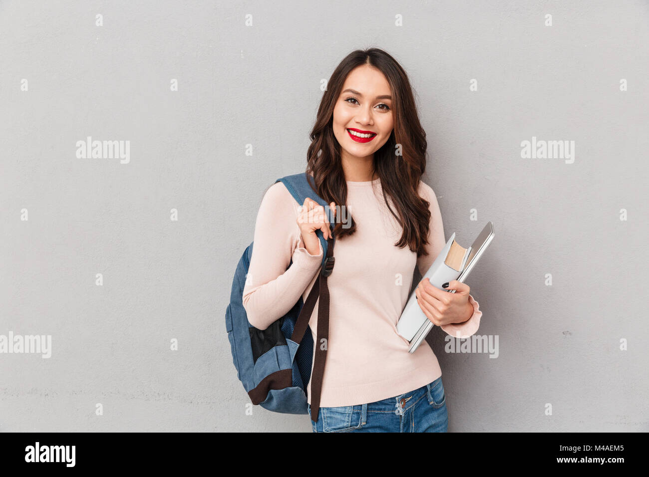 Portrait of intellectual female student with backpack being educated in university, posing with book and laptop over gray wall Stock Photo