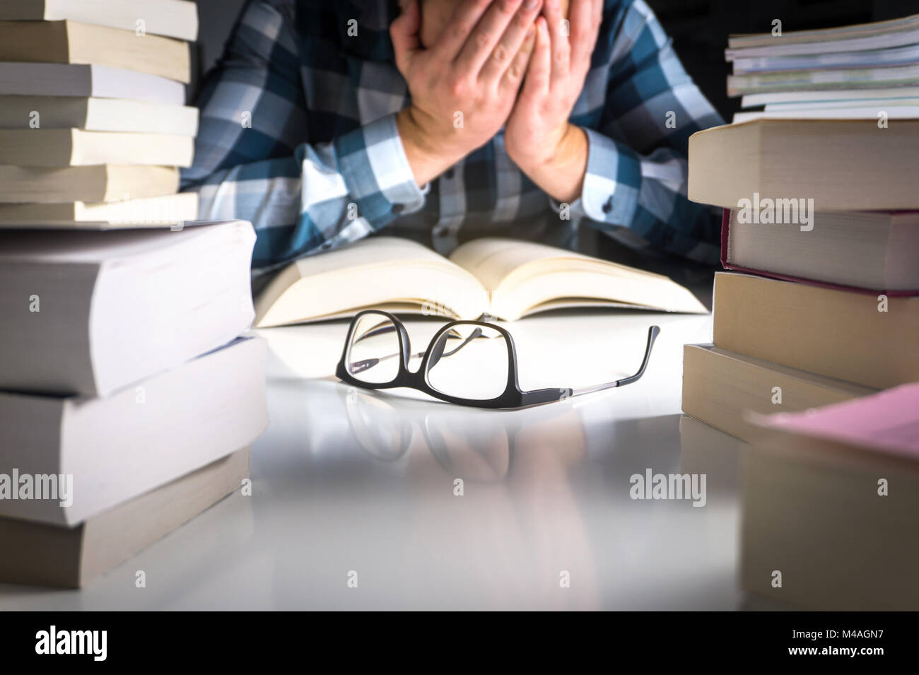 Stressed, tired and unhappy student. Too much work from school. Young man covering face with hands surrounded by stacks of books. Glasses on table. Stock Photo