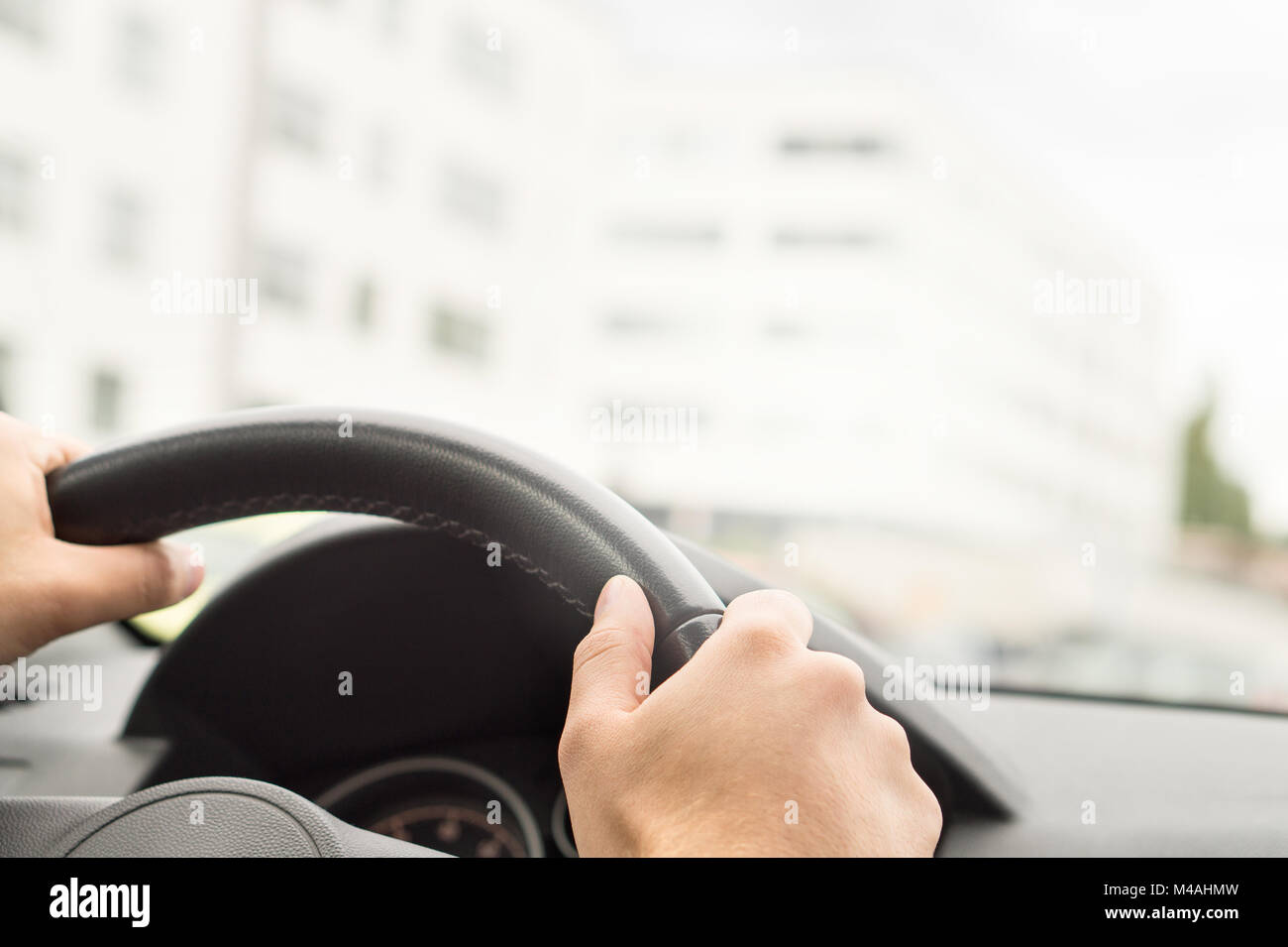 Man driving car in city. Driver holding steering wheel with both hands. Road trip, travel or commute concept. Buildings in the blurred background. Stock Photo