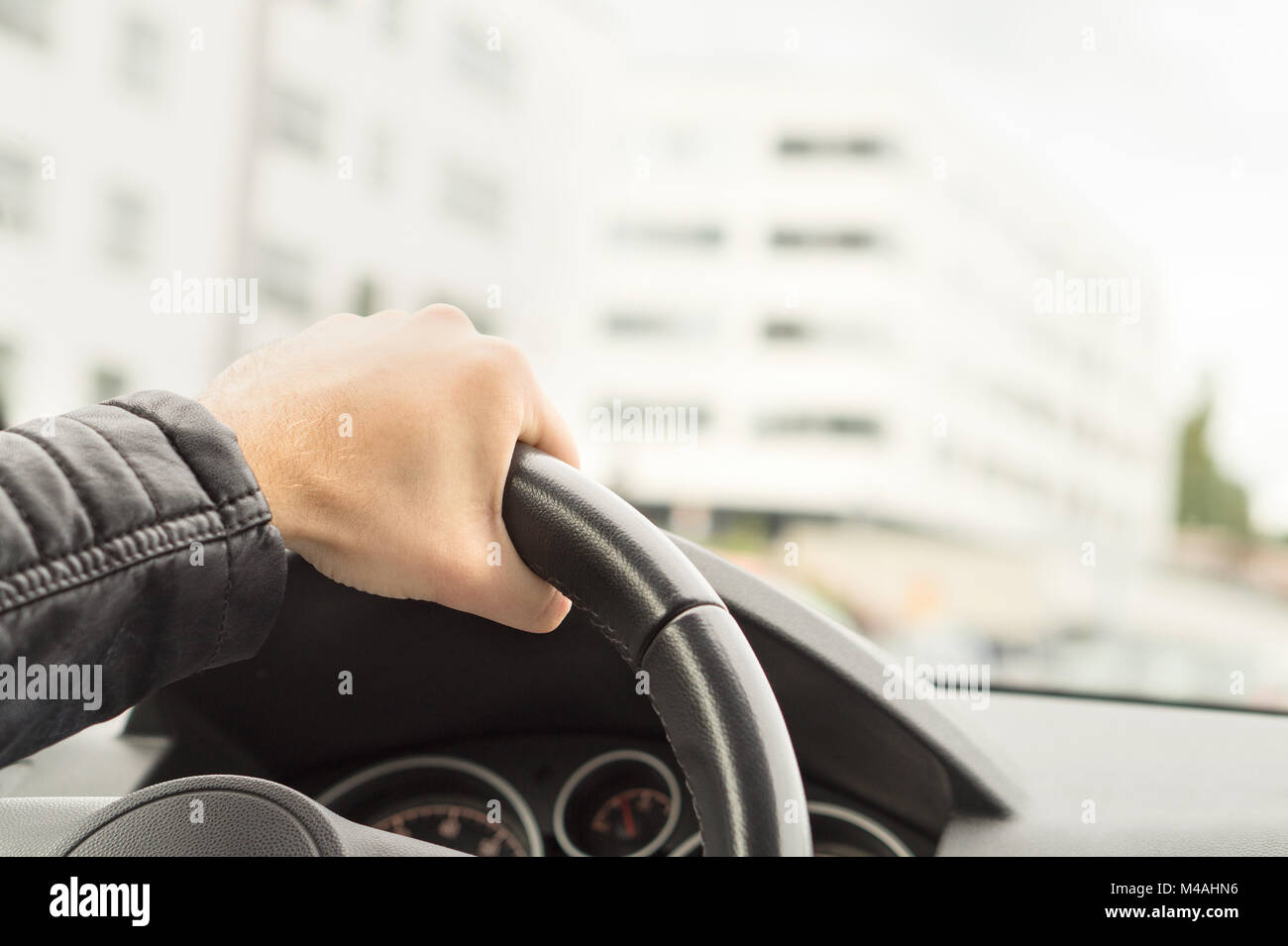 Driver holding steering wheel with one hand. Man driving car in city. Road trip, travel or commute concept. Buildings in the blurred background. Stock Photo