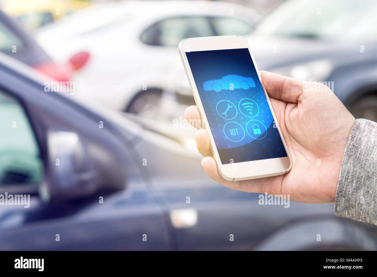 Internet of things (IOT) mobile app in smart phone for modern car. Hand holding smartphone controlling ADAS system. Vehicles parked in the background. Stock Photo