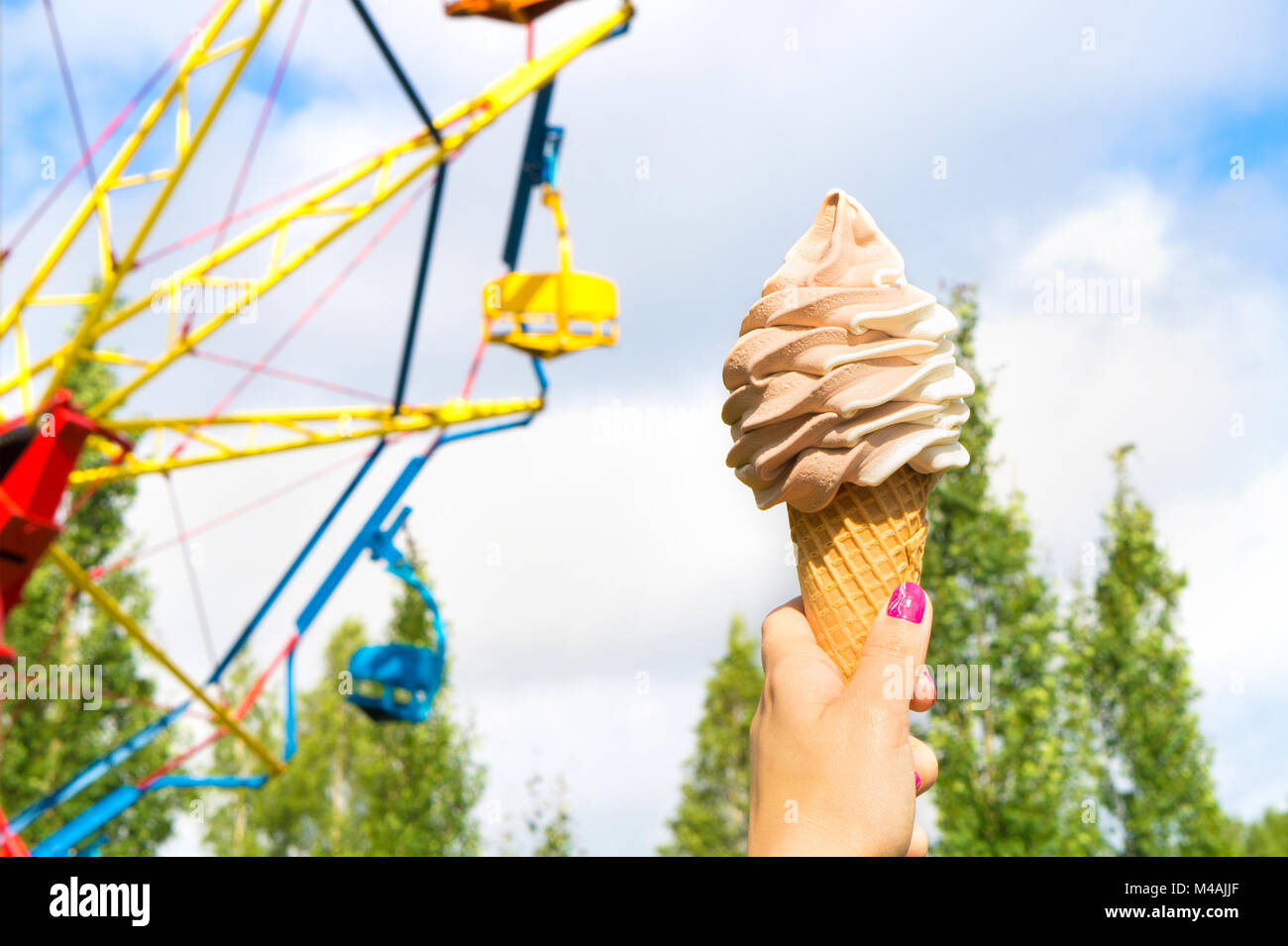 Ice cream in amusement park on a sunny summer day. Girl or young woman holding a cone of soft ice with a colorful theme park ride in the background. Stock Photo