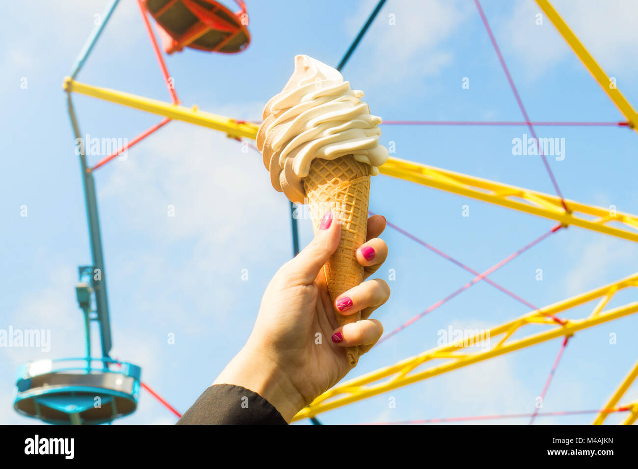 Ice cream in amusement park on a sunny summer day. Girl or young woman holding a cone of soft ice with a colorful theme park ride in the background. Stock Photo