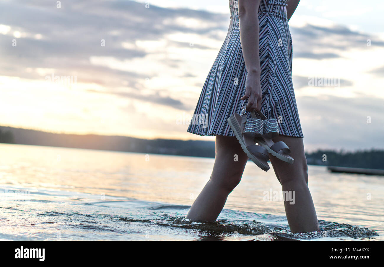 Beautiful woman in sunset walking in water and lake holding shoes in hand. Beach lifestyle view. Sad and dramatic mood. Stock Photo