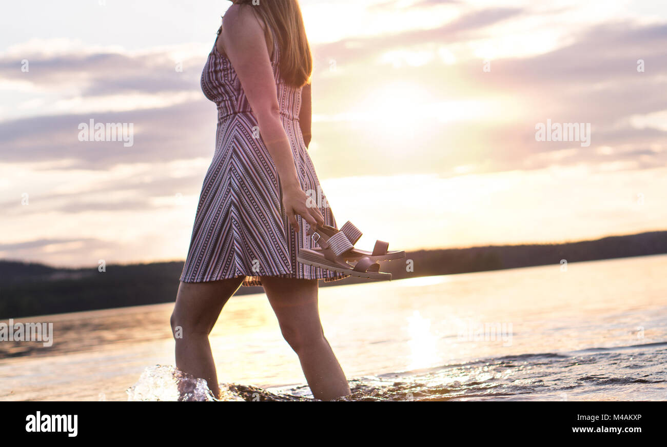 Freedom and happiness concept. Young attractive woman walking in lake water at sunset. Carefree and happy lifestyle. Holding sandals in hand. Hot summ Stock Photo