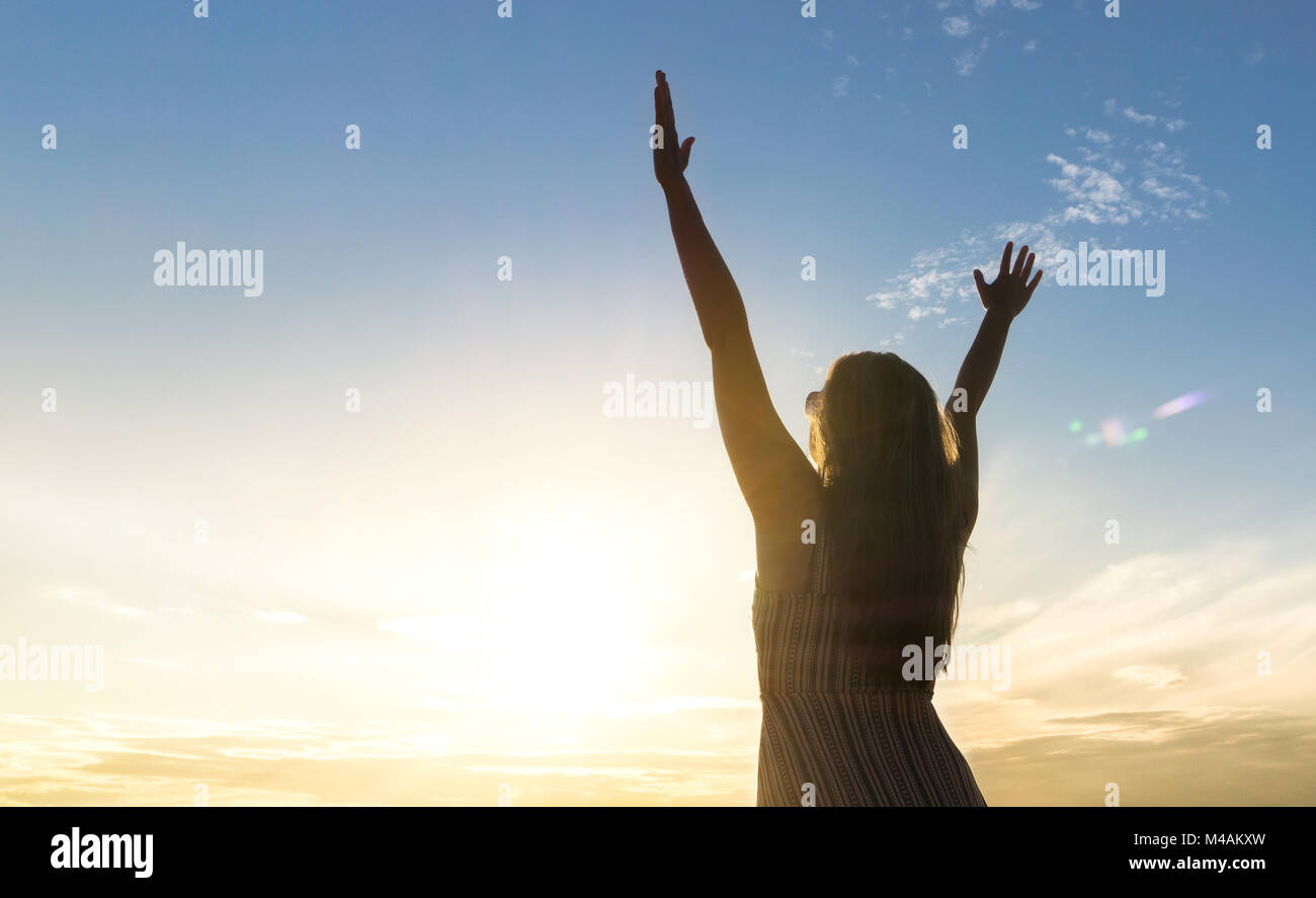 Brave happy young woman raising hands up in the air and open arms to sky. Enjoying life. Sunny healthy lifestyle concept in sunset. Stock Photo