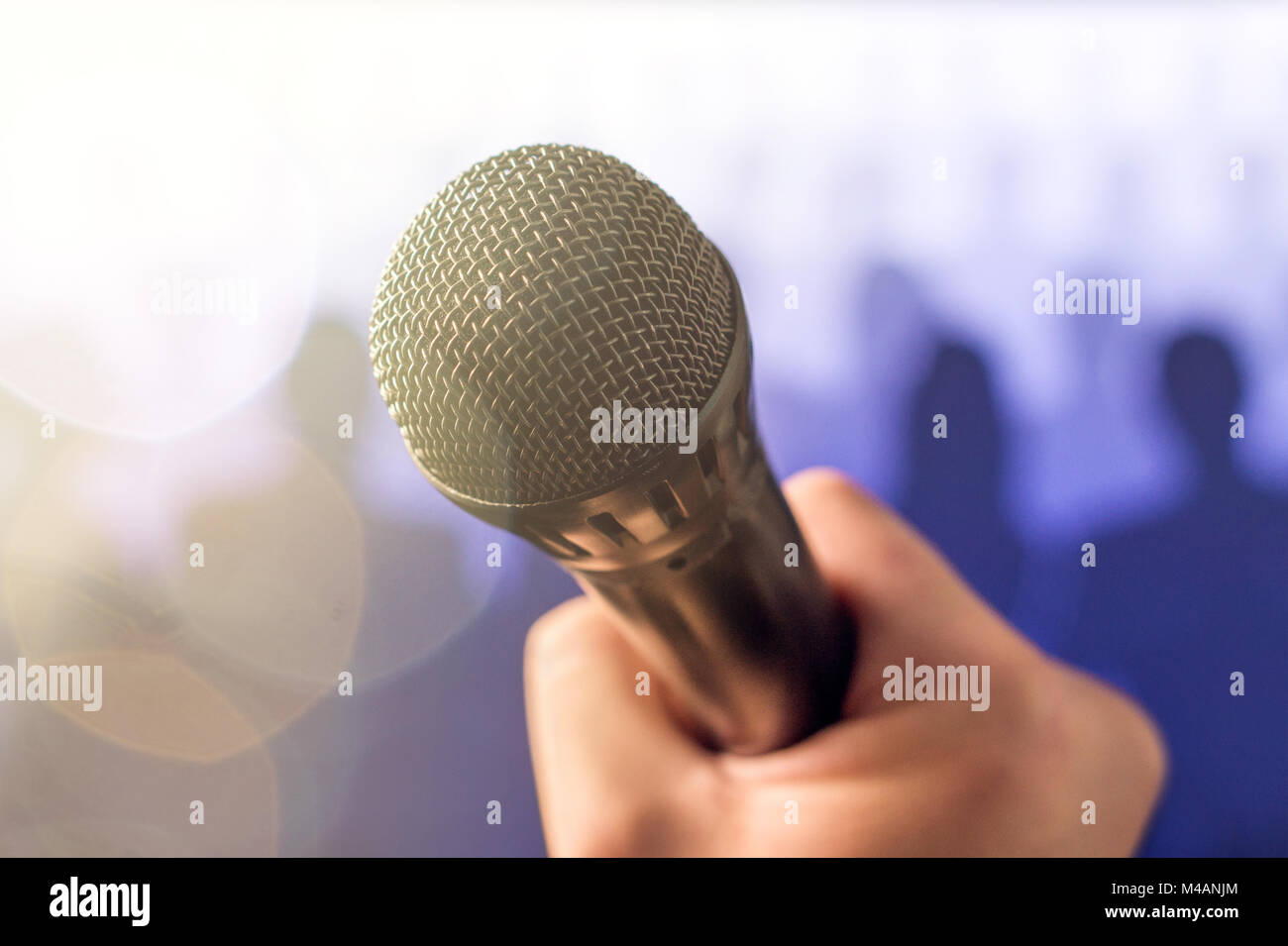 Public speaking and giving speech concept. Hand holding microphone in front of a crowd of silhouette people with lens flare and sun light leak bokeh.  Stock Photo