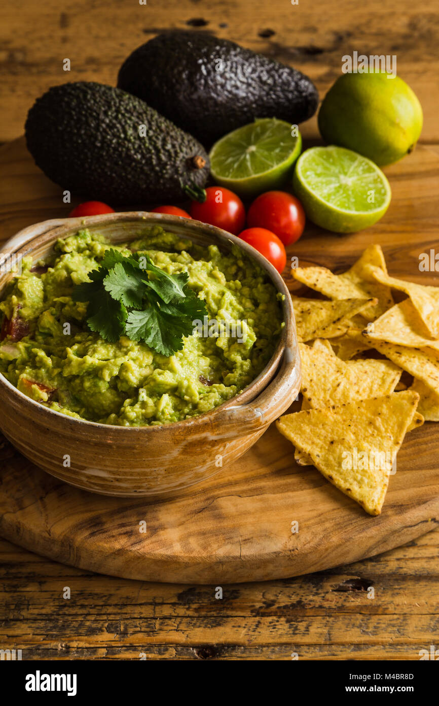 Guacamole dip in a bowl with scattered tortilla chips tomatoes avocados and limes Stock Photo