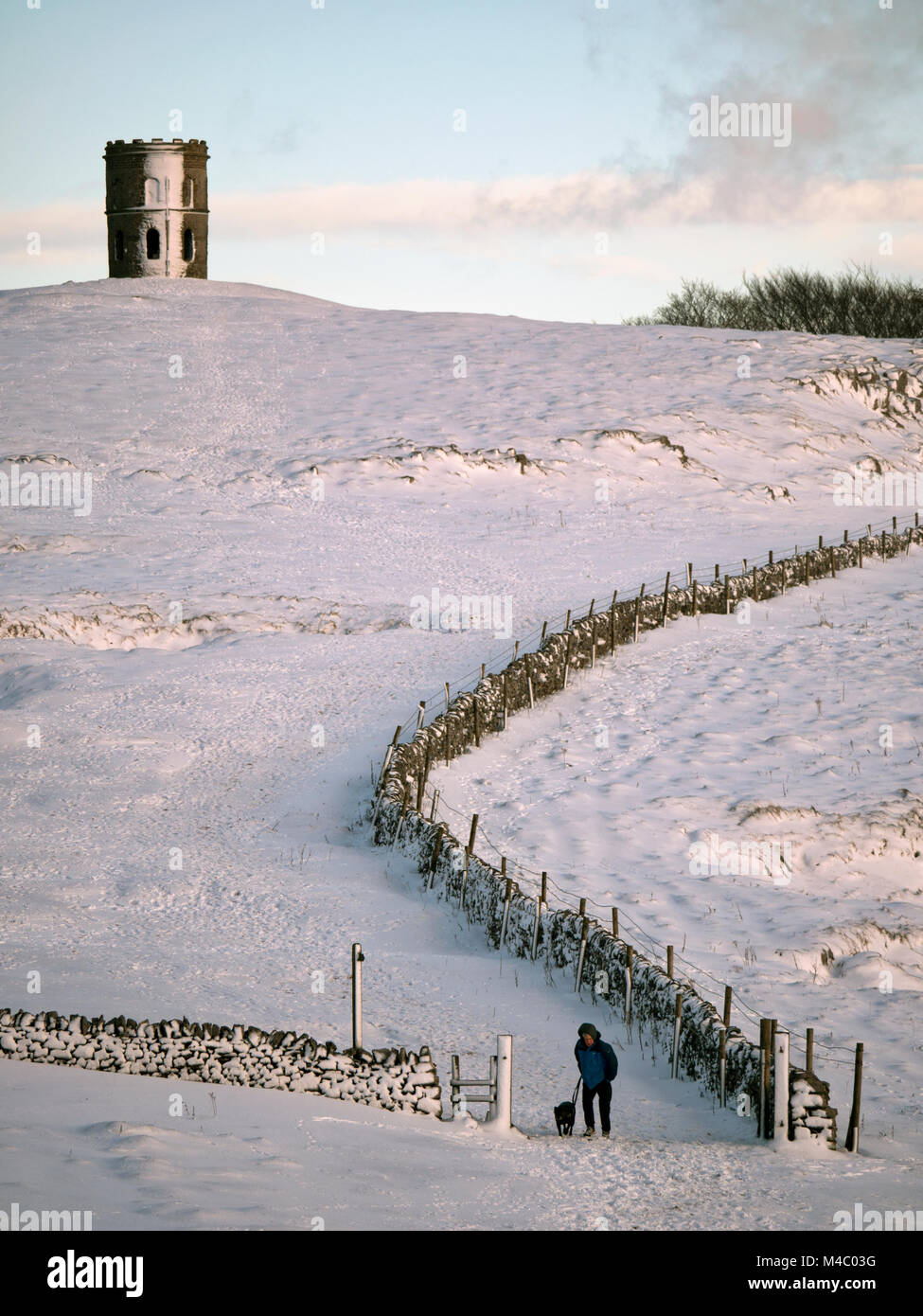 A man walking his dog in the snow near Solomon's Temple also called Grinlow Tower the Victorian Fortified hill marker above the spa town of Buxton in  Stock Photo