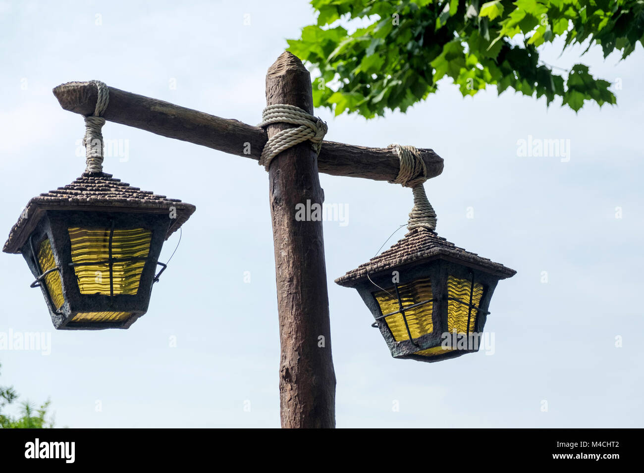 Close up of rustic wooden lamp post tied with rope & two old lamps with yellow glass hanging down.Trees on top right. -Disneyland Tokyo Stock Photo