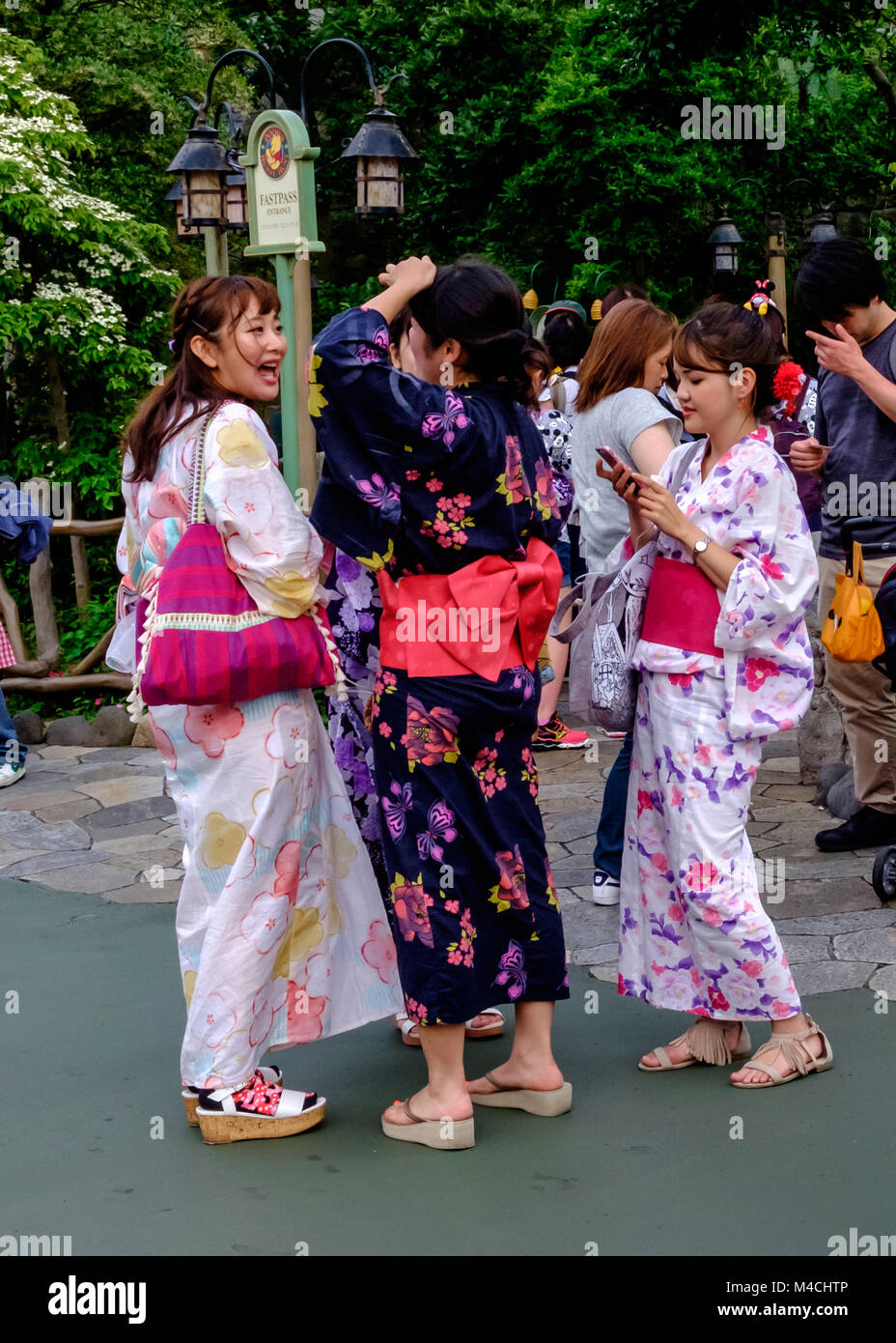 Three Japanese young girls dressed in kimonos. One is laughing, one is looking at her phone and one has her hand to her head. –At Disneyland Tokyo Stock Photo