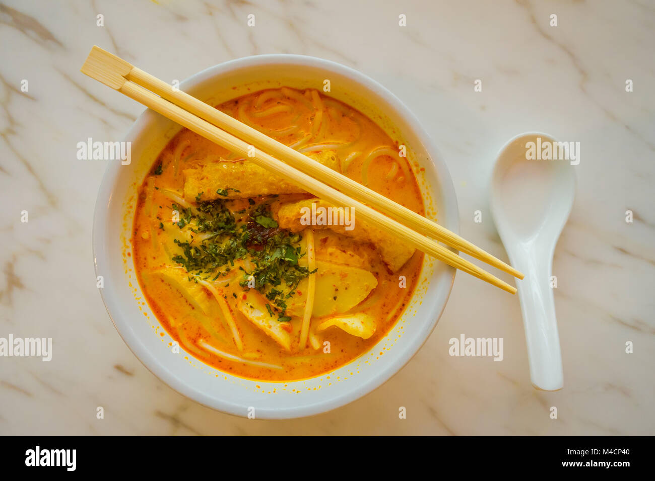 Above view of penang asam laksa rice noodle in white bowl with chopsticks on white table with spicy fish tamarind soup white porcelain spoon and fresh herbs in Singapore Stock Photo
