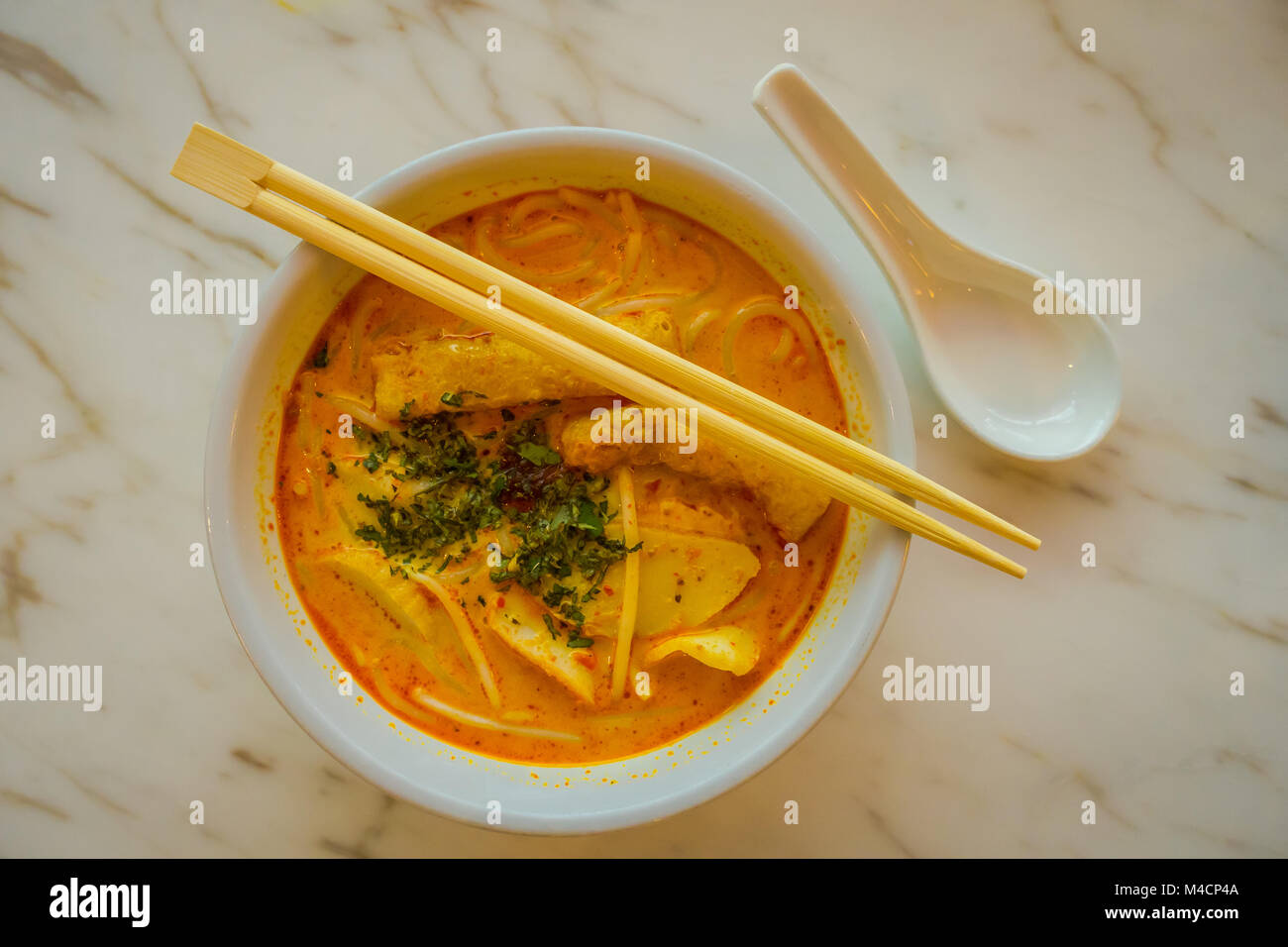 Above view of penang asam laksa rice noodle in white bowl with chopsticks on white table with spicy fish tamarind soup white porcelain spoon and fresh herbs in Singapore Stock Photo