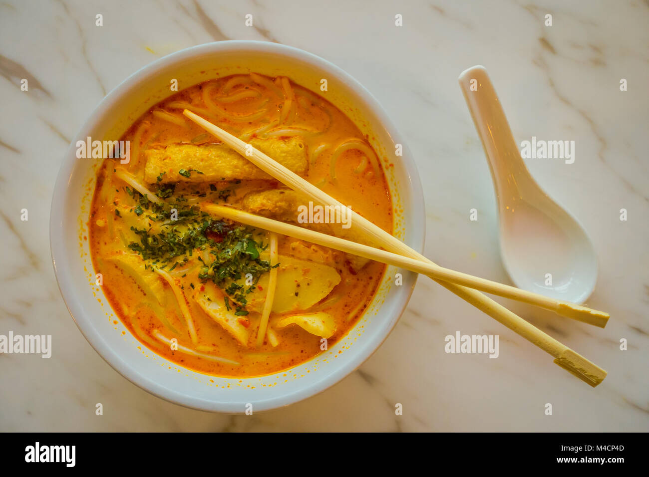 Above view of penang asam laksa rice noodle in white bowl with chopsticks inside the bowl on white table with spicy fish tamarind soup and white porcelain spoon and fresh herbs in Singapore Stock Photo