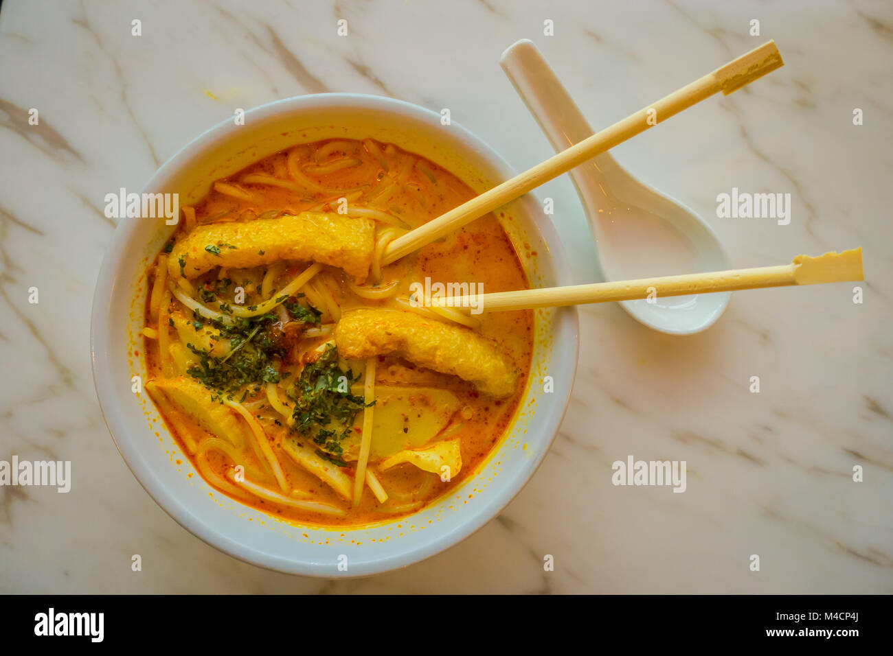 Above view of penang asam laksa rice noodle in white bowl with chopsticks inside the bowl on white table with spicy fish tamarind soup and white porcelain spoo and fresh herbs in Singapore Stock Photo