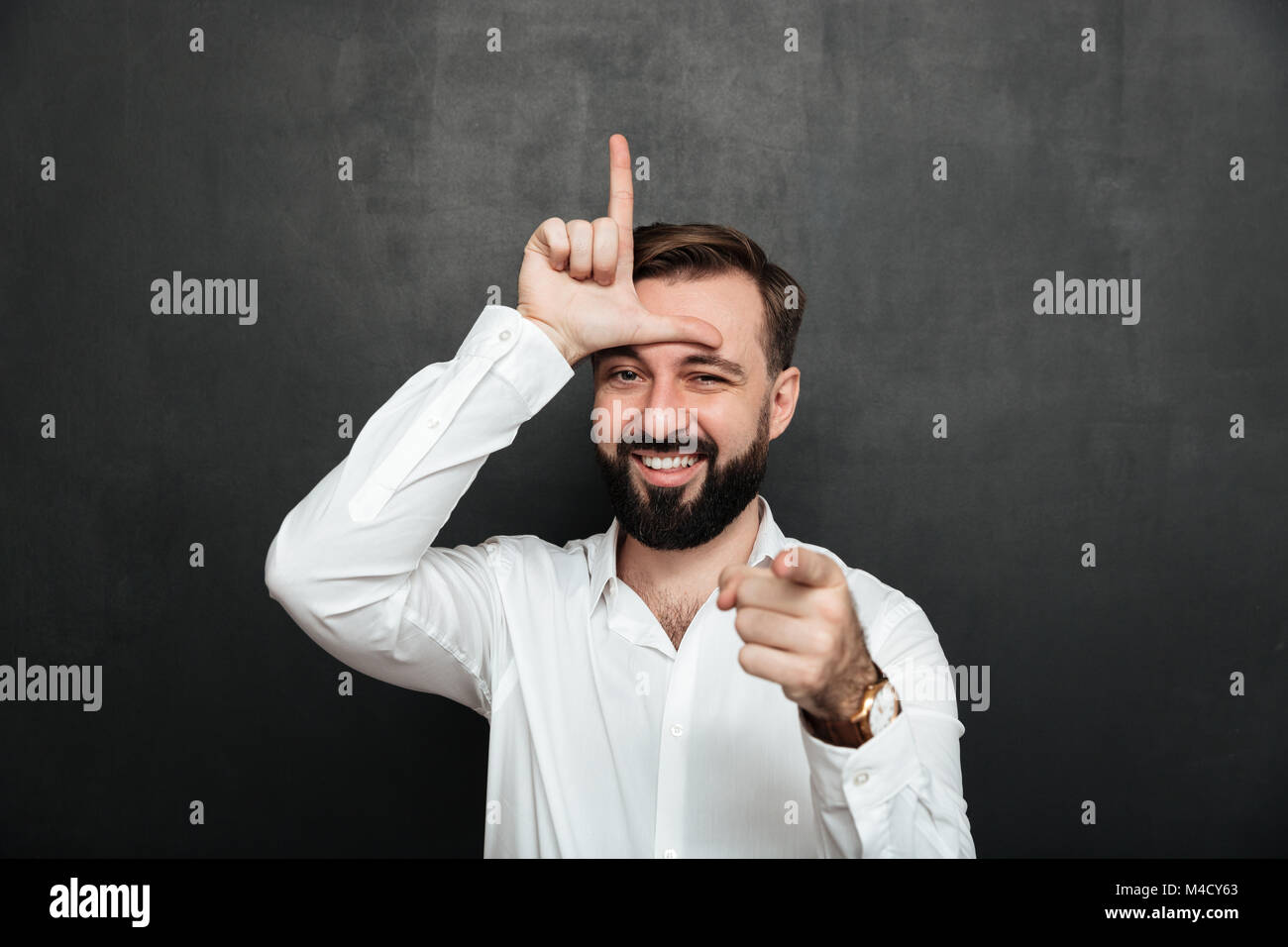 Portrait of sarcastic man showing loser sign on his forehead and pointing on camera with smile mocking or humiliate over graphite wall Stock Photo