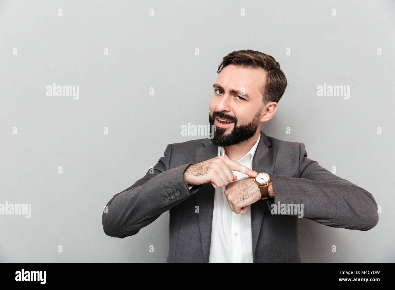 Horizontal image of bearded man pointing at his wrist watch posing isolated over gray background Stock Photo