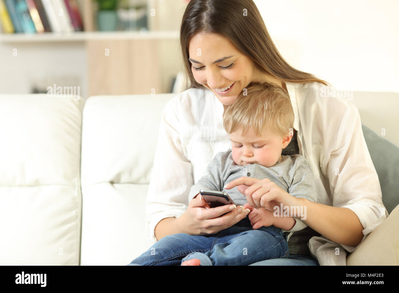 Mother showing smart phone content to his baby son sitting on a couch in the living room at home Stock Photo
