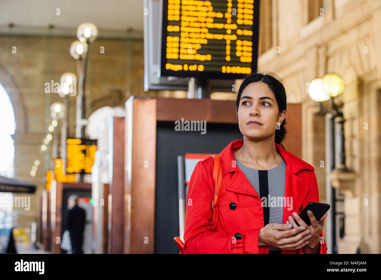 Businesswoman is using her smart phone to find her train time using the departure board. Stock Photo