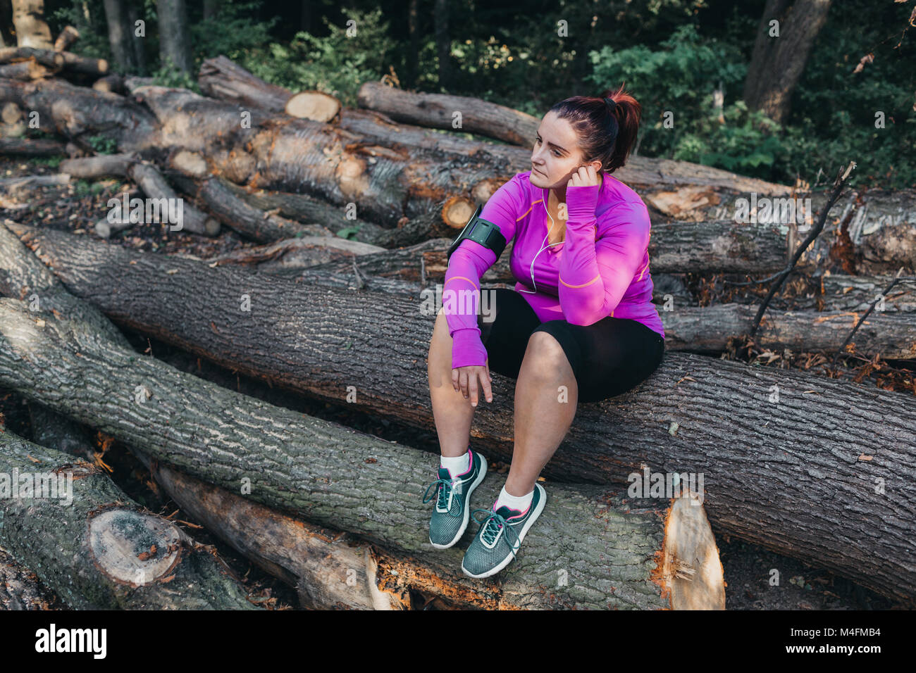 Portrait of a woman with earphones sitting on a pile of logs and resting in the nature. Stock Photo