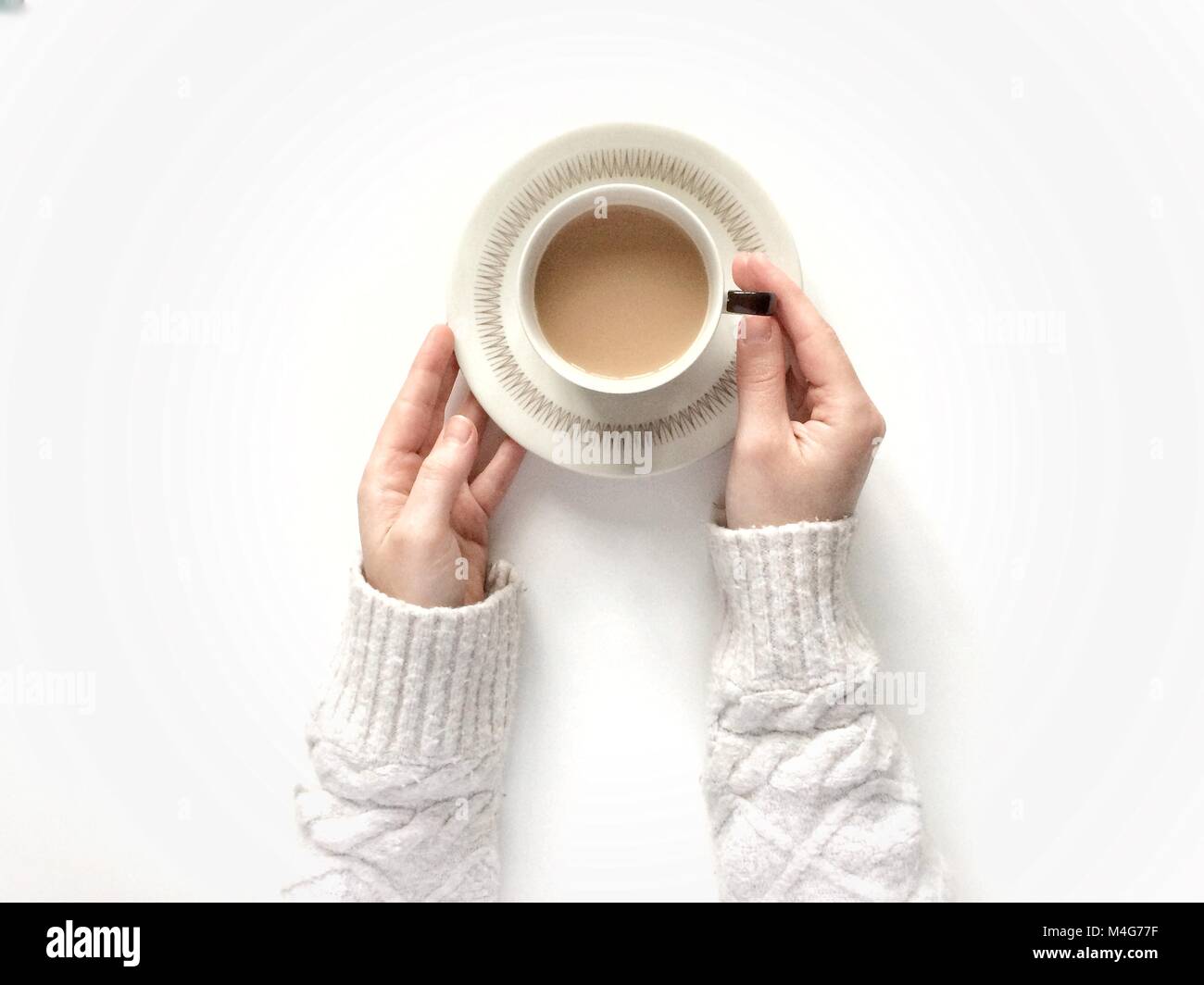 Looking down from above onto a white background with a girl's arms holding a cup of tea in a cup and saucer whilst wearing a cosy and warm swaeater. Stock Photo