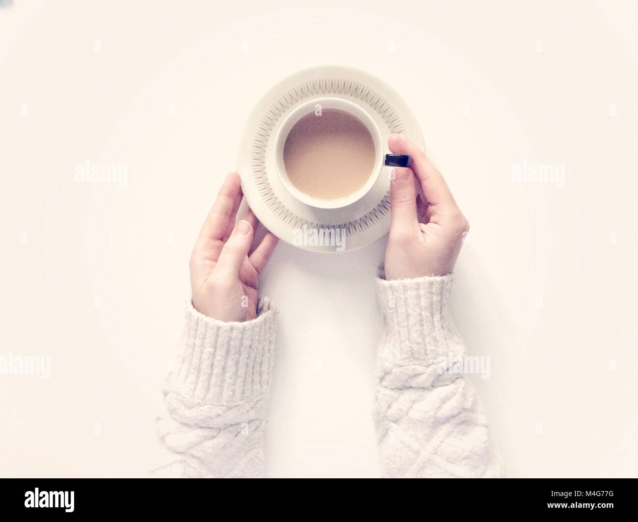 Looking down from above onto a white background with a girl's arms holding a cup of tea in a cup and saucer whilst wearing a cosy and warm swaeater. Stock Photo