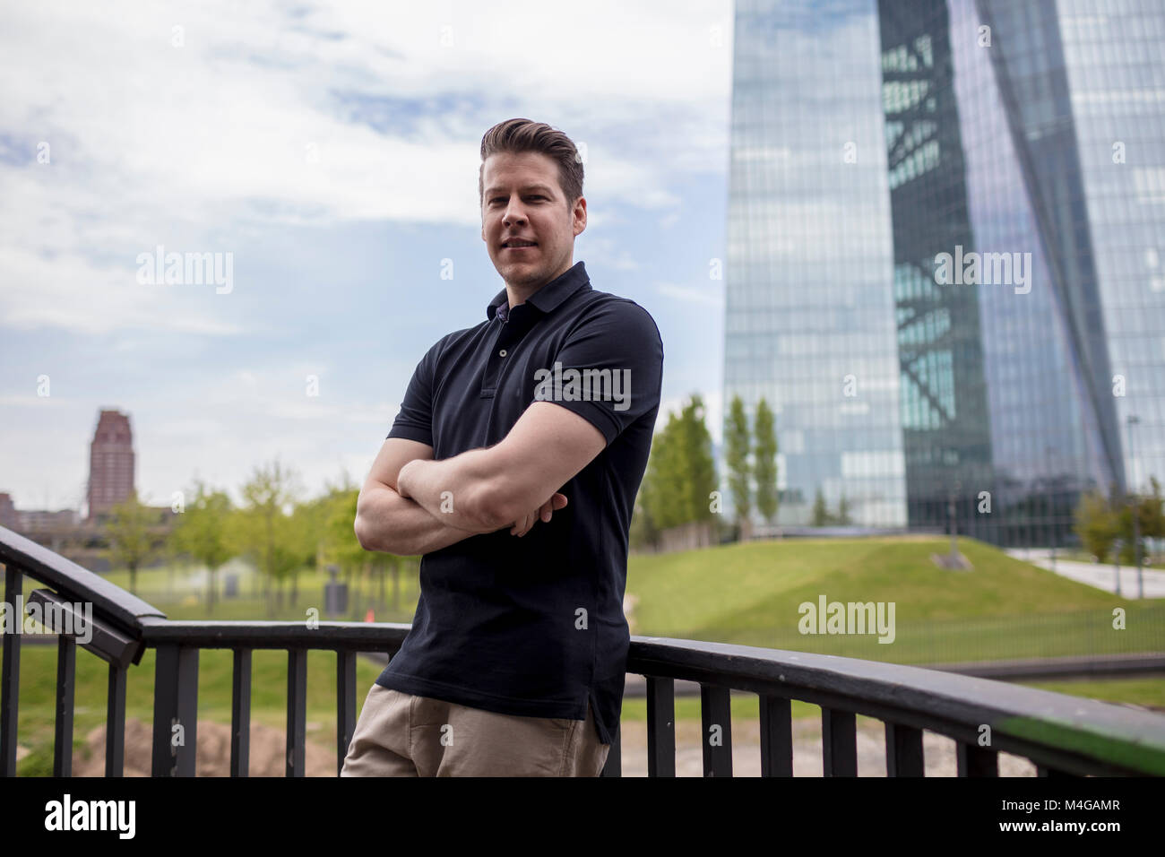 A man standing with his arms crossed leaning on a balustrade and a skyscraper building in the background. Stock Photo