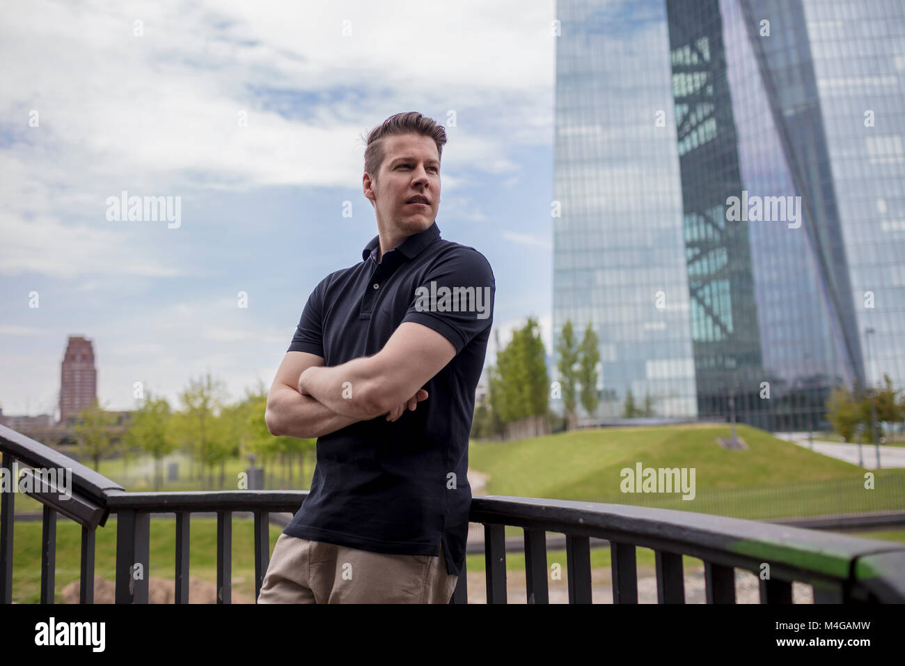 A man standing with his arms crossed leaning on a balustrade and a skyscraper building in the background. Stock Photo