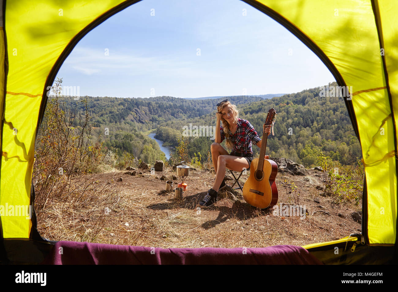 Young blonde woman tourists with guitar in camp on cliff over river and forest landscape. View from camping tent entrance. Stock Photo