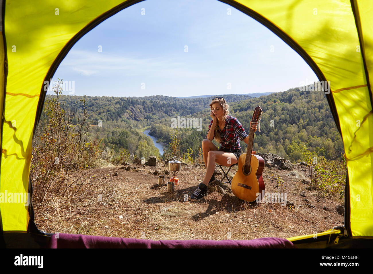 Young blonde woman tourists with guitar in camp on cliff over river and forest landscape. View from camping tent entrance. Stock Photo