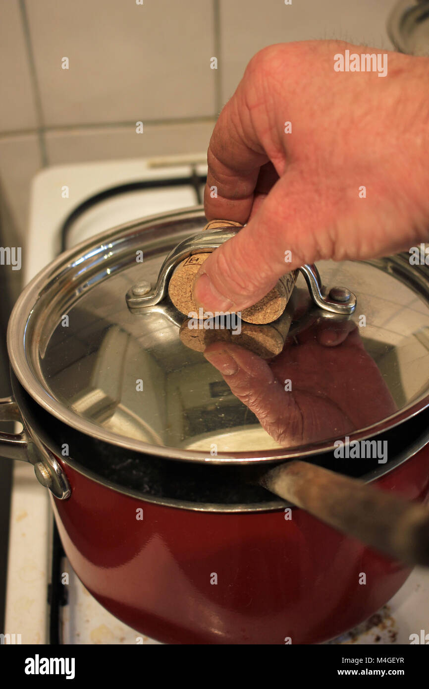 A cook lifts a hot metal lid off a pan. He has put two used wine bottle corks under the pan