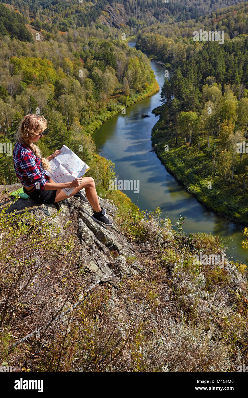 Young blonde woman tourist  sitting on a cliff with map in hands on background of siberian autumn landscape with the river Berd. Russia, Siberia, Sala Stock Photo