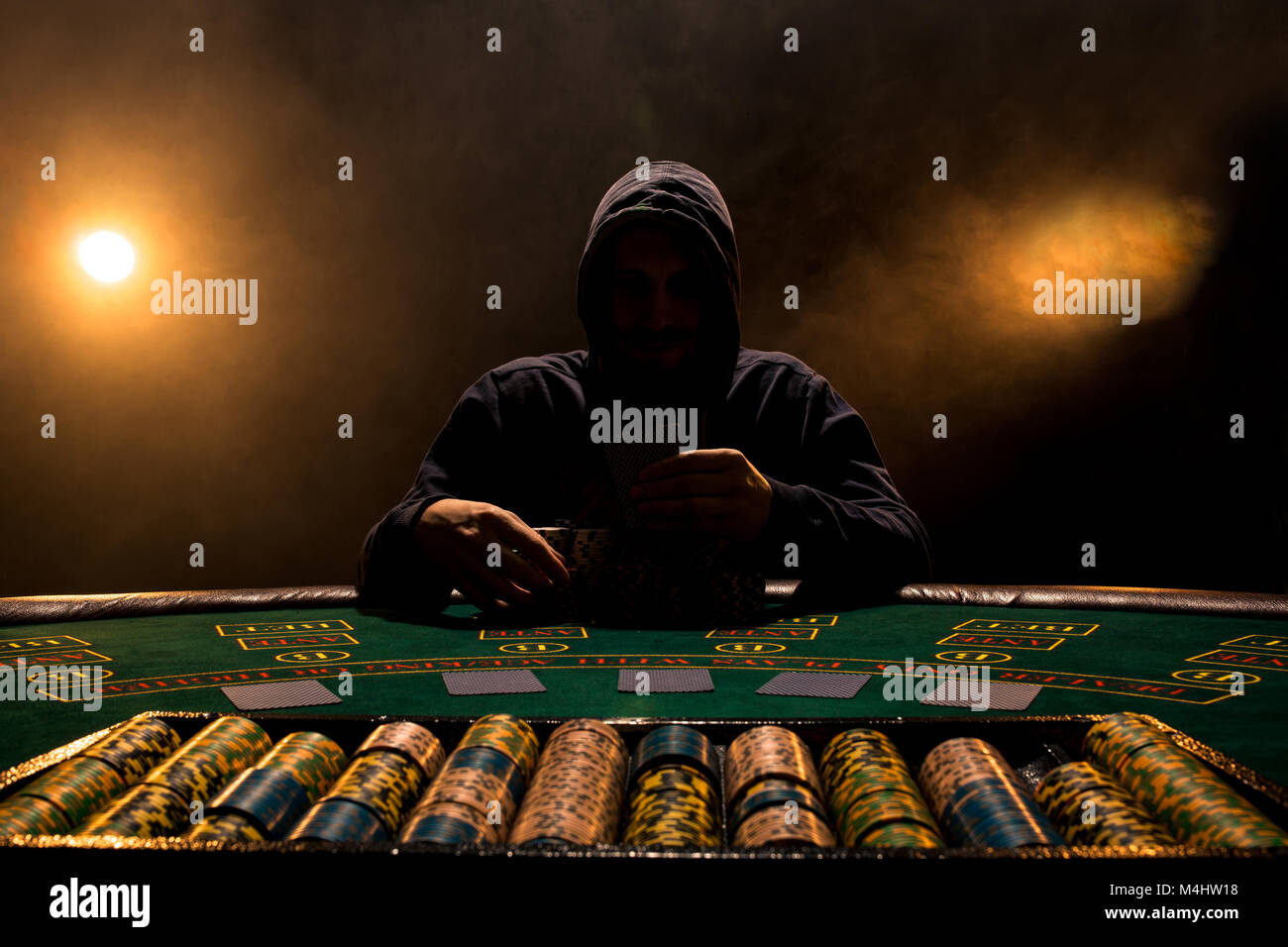 Portrait of a professional poker player sitting at poker table. A man in a  hood with cards in his hands sits at a poker table in a dark room full of c
