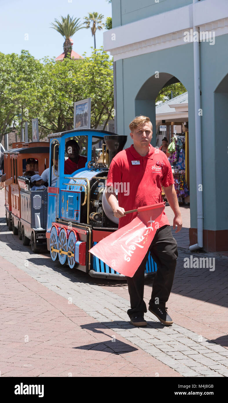 Young man carrying a red flag walks in front of a children's train ride. Waterfront, Cape Town, South Africa, Stock Photo