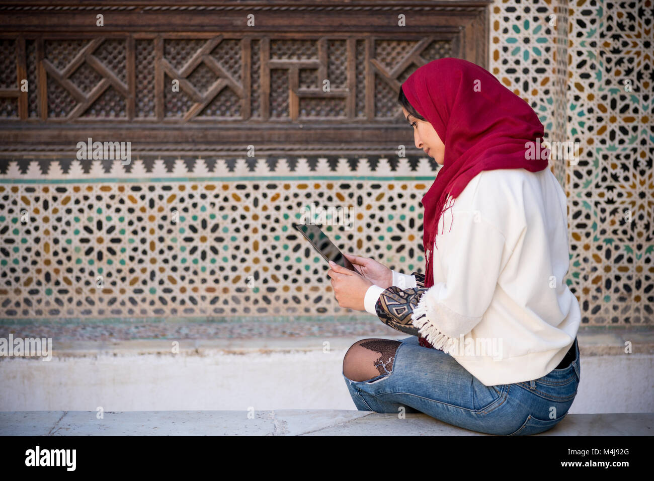 Muslim woman working on tablet in traditional clothing with red headscarf on her head beside a traditional arabesque Moroccan wall Stock Photo