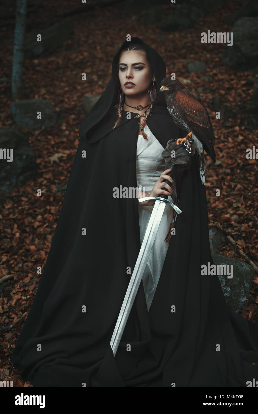 Proud hooded woman with sword and hawk. Forest background Stock Photo