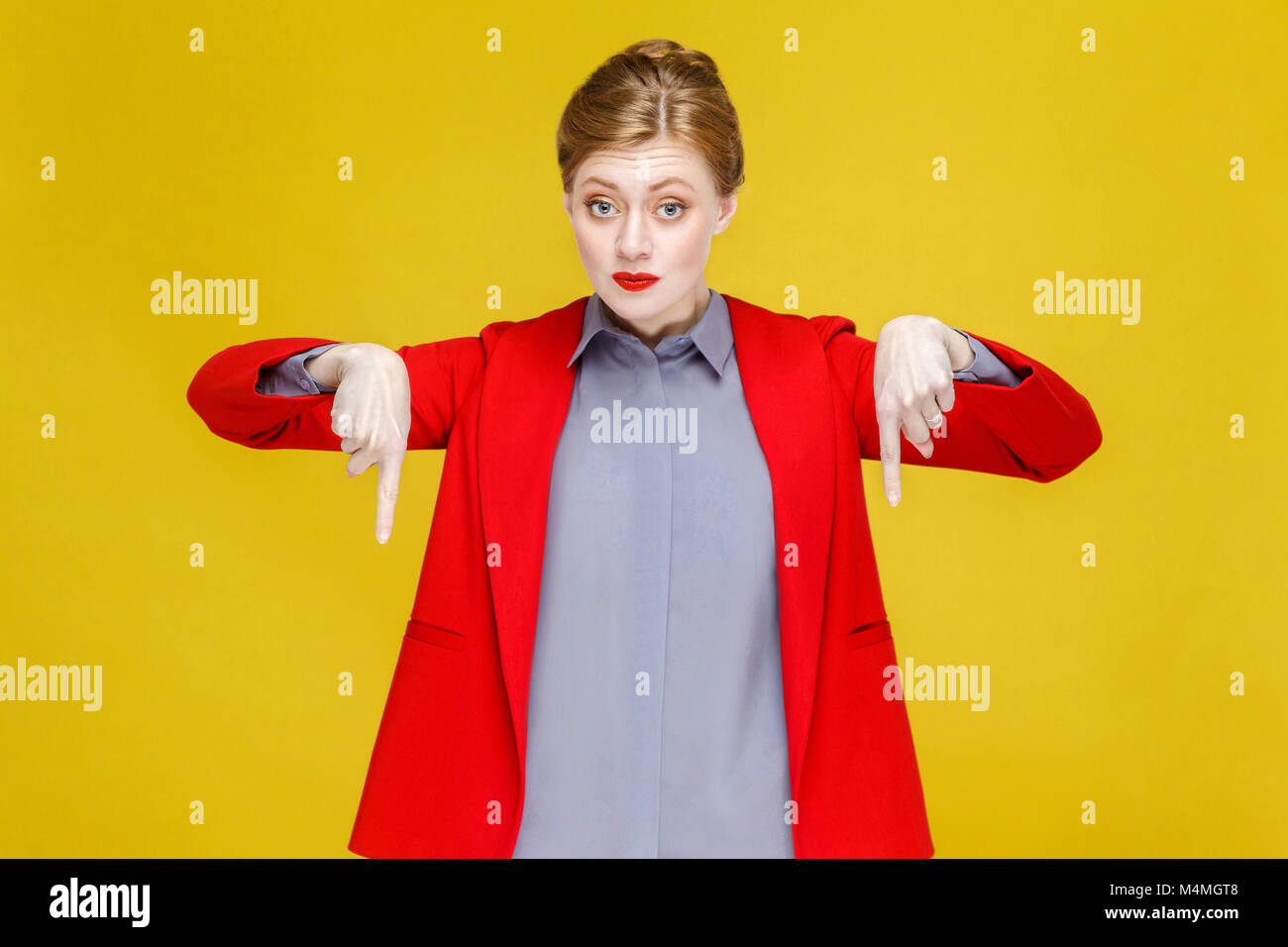 Ginger red head woman in red suit wish pointing down at copy space. Studio shot, isolated on yellow background Stock Photo